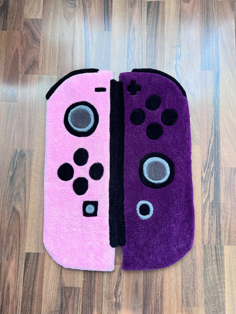 May include: A pink and purple rug shaped like a Nintendo Switch game console. The rug is made of soft, plush material and features a detailed design of the console's buttons and controls.