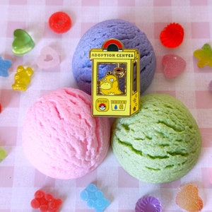 May include: A yellow enamel pin featuring a cartoon Pokemon character in a vending machine with the text "Adoption Center". The pin is on a pink and white checkered background with three scoops of ice cream in the background.