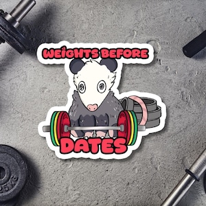 May include: A sticker featuring a cartoon opossum lifting weights. The opossum is gray and white, with large eyes and a pink nose. The barbell has red, green, and yellow weights. Text reads "WEIGHTS BEFORE DATES" in a red and white font.