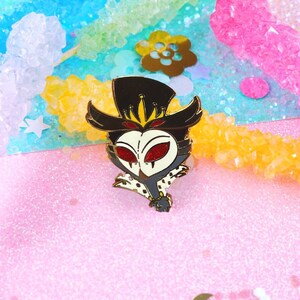 May include: A black and white enamel pin featuring a cartoon character with a crown, a top hat, and red eyes. The character is wearing a black and white patterned coat and has a gold chain around its neck.