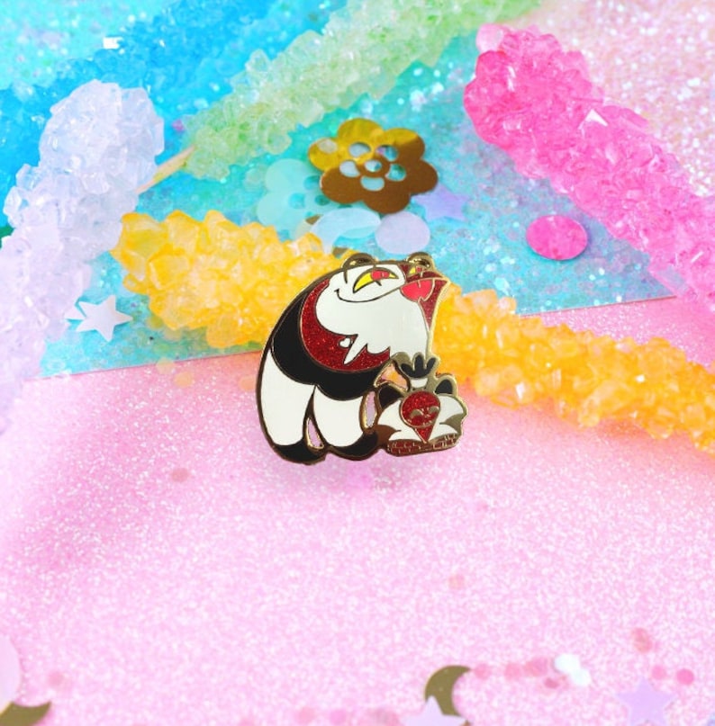 May include: Gold enamel pin featuring a white and black cartoon cat with red eyes and a red heart in its paw. The cat is wearing a black and red collar.