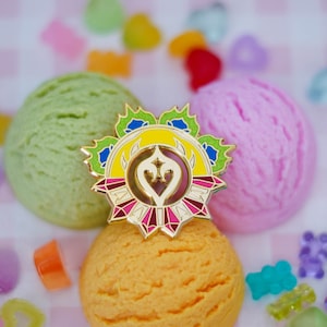 Könnte beinhalten: Goldene Emaille-Anstecknadel mit einem gelben, blauen und grünen Design. Die Anstecknadel zeigt eine stilisierte Herzform mit einem Flammenmuster. Die Anstecknadel ist von einem Ring aus rosa und roten Edelsteinen umgeben. Die Anstecknadel liegt auf drei Kugeln Eis: grün, rosa und orange.