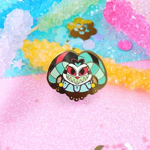 May include: A gold-colored enamel pin featuring a cartoon character with red, black, and blue details. The character has a large, toothy grin and a heart-shaped design on its chest. The pin is set against a pink, sparkly background.