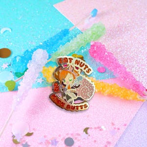 Può includere: Una spilla smaltata colorata con un personaggio dei cartoni animati con i capelli arancioni e uno scoiattolo. La spilla ha il testo "EAT NUTS" e "RICH BUTTS". La spilla è circondata da caramelle dure colorate su uno sfondo glitterato.