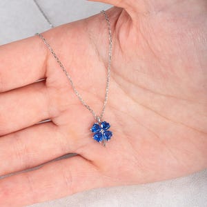 Könnte beinhalten: Eine silberfarbene Halskette mit einem vierblättrigen Kleeblatt-Anhänger. Das Kleeblatt besteht aus vier herzförmigen, leuchtend blauen Edelsteinen in einer Metallfassung und hängt an einer zarten, dünnen silberfarbenen Kette mit Gliedern.