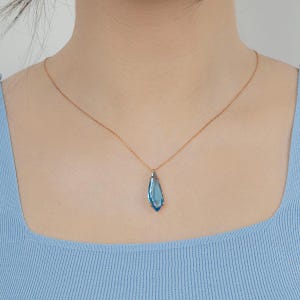 Può includere: Una collana color oro con un pendente a goccia in pietra preziosa blu chiaro. Il pendente è sospeso a una delicata catena. La collana è indossata su un top azzurro.
