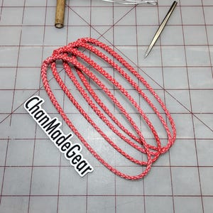 Puede incluir: Una colección de bucles de cordón trenzado rojo, un mango de madera con alambre, una lezna de metal y un calibrador están dispuestos sobre una superficie de cuadrícula. El texto "ChanMadeGear" es visible. Esta imagen probablemente esté relacionada con la artesanía o la marroquinería.