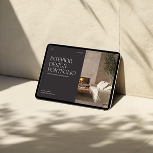 Op de afbeelding: Een tabletcomputer die een website voor een interieurontwerp portfolio weergeeft. De website heeft een minimalistisch ontwerp met de tekst "INTERIOR DESIGN PORTFOLIO" in wit op een zwarte achtergrond. De website toont ook een foto van een woonkamer met een open haard en een bank.