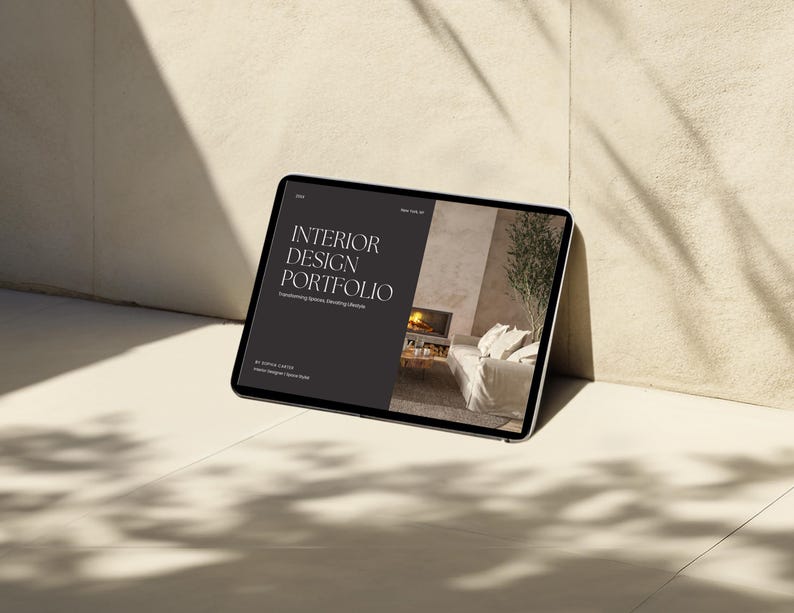 Op de afbeelding: Een tabletcomputer die een website voor een interieurontwerp portfolio weergeeft. De website heeft een minimalistisch ontwerp met de tekst "INTERIOR DESIGN PORTFOLIO" in wit op een zwarte achtergrond. De website toont ook een foto van een woonkamer met een open haard en een bank.