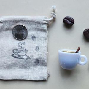 May include: A white ceramic coffee cup with a brown cinnamon stick, a small bag with a coffee cup design, and three brown coffee beans on a white background.