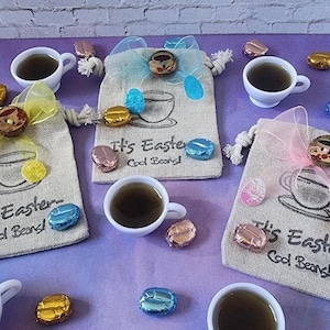 May include: Three small burlap bags with a light blue, pink, and yellow ribbon tied around the top. Each bag has a white cup and saucer design with the text "It's Easter Cool Beans!" printed on the front. The bags are filled with chocolate candies.