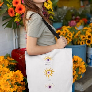 May include: White tote bag with a design of three stylized eyes with sun rays and the text "Protect Your Energy".