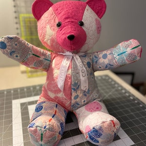 May include: A pink and white patchwork teddy bear with a pink bow tied around its neck. The bear is sitting on a gray and white cutting mat. The bow has the text "COOTIE" printed on it.