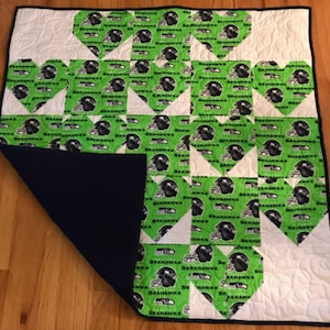 May include: A green and white quilt with a Seattle Seahawks theme. The quilt is made of squares with the Seahawks logo and the words "Seahawks" repeated on each square. The quilt is backed with a dark blue fabric.