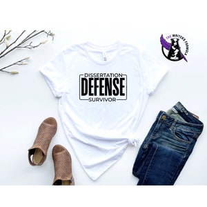 May include: White t-shirt with black text that reads "Dissertation Defense Survivor". The shirt is laying on a white surface with a pair of brown sandals and blue jeans.