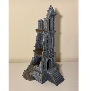 May include: A gray stone tower ruin with a broken top and a doorway at the bottom. The tower is made of stone blocks and has a weathered look. The tower is on a white surface.