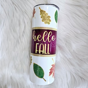 May include: A white tumbler with a black lid, featuring a burgundy band with the words "hello FALL" in gold script. The tumbler is decorated with gold glitter and leaf designs in green, gold, and red.
