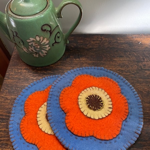 Puede incluir: Dos posavasos redondos de fieltro azul con diseños de flores naranjas y costuras marrones. Una tetera verde con un diseño floral se encuentra en el fondo.