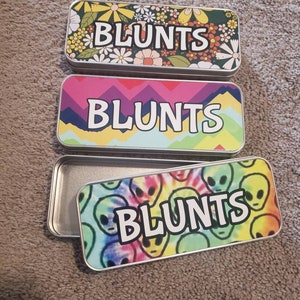 May include: Three rectangular metal tins with the word "BLUNTS" in white block letters. The top tin has a floral design with yellow, orange, and white flowers on a dark green background. The middle tin has a pink, purple, and blue geometric design. The bottom tin has a tie-dye alien design.