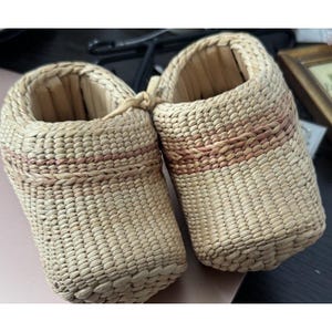 May include: Two woven straw baby shoes. The shoes are light tan with a reddish-brown band around the middle. The shoes are tied together with a woven cord.