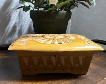May include: A small, rectangular wooden box with a light yellow finish. The lid features a carved sunburst design, while the sides are decorated with carved geometric shapes. The box is likely used for storing small items.