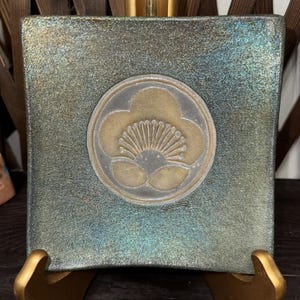 May include: Square decorative plate with a metallic finish in shades of green and gold. The plate features a central emblem of a stylized flower in gold and silver. The plate is displayed on a gold-colored stand.