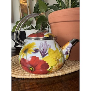 May include: White enamel teapot with a floral design featuring red poppies, yellow daffodils, and purple irises. The teapot has a silver handle and spout, and a red and silver lid. The teapot is sitting on a woven mat.