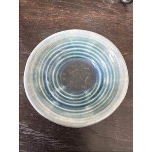 May include: A handmade ceramic bowl with concentric blue and teal rings. The center of the bowl is a darker shade of blue with brown accents. The bowl has a slightly uneven rim and a rustic aesthetic.