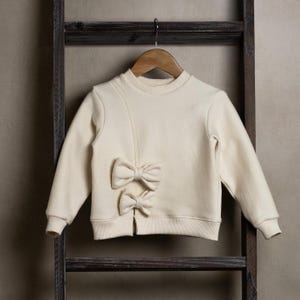 Może przedstawiać: Kremowa bluza z kokardką z tyłu. Bluza wisi na drewnianej drabinie.