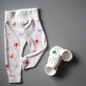 May include: White baby leggings with a colorful animal print design. The leggings have a white elastic waistband. Two white plastic clogs with a green clover design are also pictured.