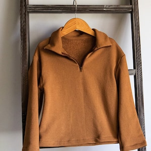 Könnte beinhalten: Ein brauner, langärmliger Pullover mit 1/4-Reißverschluss, der an einem Holzbügel hängt.