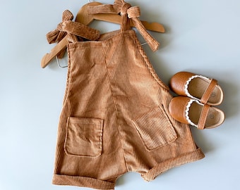 May include: A brown corduroy overall with a tie at the shoulders. The overall has two pockets and is hanging on a wooden hanger. There are two brown leather shoes with white trim on the side.
