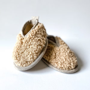 Op de afbeelding: Een paar beige wollen pantoffels met een zachte, pluche textuur. De pantoffels zijn gemaakt van een comfortabel materiaal en hebben een eenvoudig ontwerp.