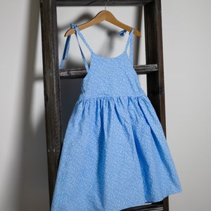 以下が含まれることがあります： 白い花柄の薄い青色のノースリーブドレス。ドレスはギャザーの入ったスカートと、肩で結ぶ細いストラップが付いています。