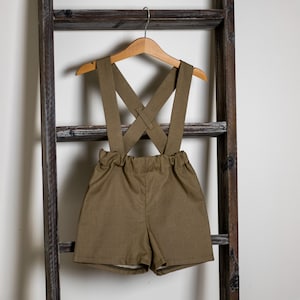 Può includere: Shorts di colore verde oliva con bretelle regolabili. Le bretelle sono incrociate sul retro e hanno una chiusura a bottone.