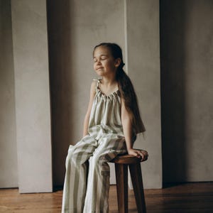 May include: A young girl wearing a green and white striped jumpsuit sits on a wooden stool. She has her eyes closed and is smiling.
