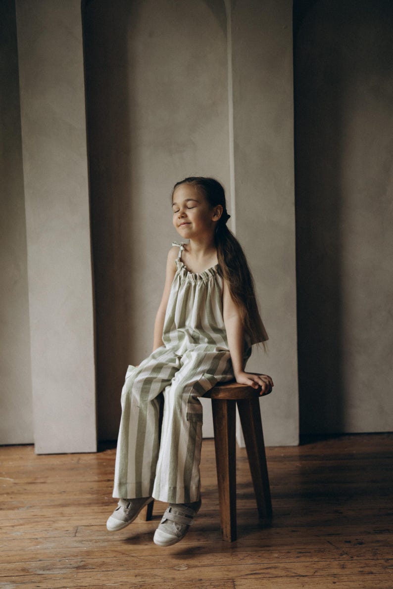 May include: A young girl wearing a green and white striped jumpsuit sits on a wooden stool. She has her eyes closed and is smiling.