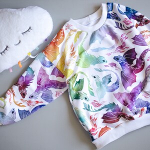 Puede incluir: Sudadera blanca con estampado de drag&oacute;n de acuarela arco&iacute;ris. Los dragones est&aacute;n volando y tienen llamas saliendo de sus bocas.