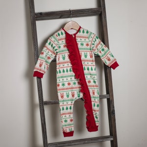Könnte beinhalten: Ein cremefarbener Baby-Einteiler mit einem Weihnachtsmuster aus Rentieren, Bäumen und Schneeflocken in Rot und Grün. Der Einteiler hat eine rote Rüsche vorne und rote Bündchen. Er hängt an einem Holzbügel.