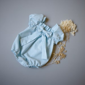 May include: A light blue baby onesie with a bow at the neckline. The onesie has short sleeves and a ruffled neckline.