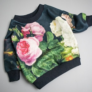 Puede incluir: Una sudadera azul oscuro con estampado floral de rosas rosas, blancas y verdes. La sudadera tiene cuello redondo y mangas largas.