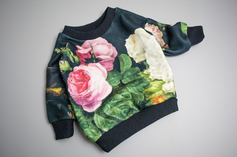 Puede incluir: Una sudadera azul oscuro con estampado floral de rosas rosas, blancas y verdes. La sudadera tiene cuello redondo y mangas largas.
