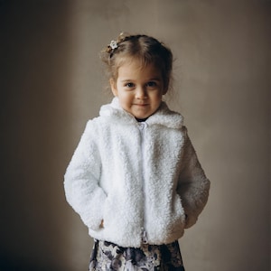 Peut inclure: Une jeune enfant portant une veste blanche à capuche, moelleuse et zippée. La veste a un aspect doux et texturé. L'enfant porte également une jupe à motifs floraux. Un petit accessoire de cheveux en forme de fleur est visible.