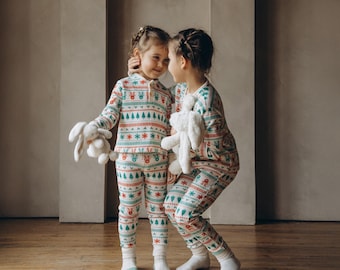 May include: Two children wearing matching Christmas pajamas with a green, red, and white pattern. They are holding stuffed animals and looking at each other.