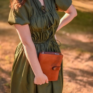 May include: A woman wearing a green dress holds a brown leather journal with an owl design. The journal has a metal clasp and a strap.
