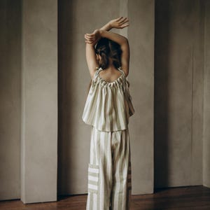May include: A girl wearing a green and white striped top and matching pants. The outfit is made of a soft, lightweight fabric. The girl is standing in front of a wall with arched doorways.