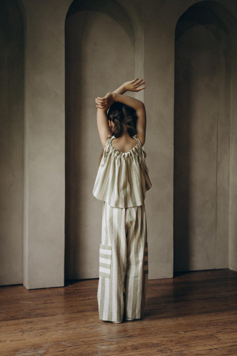 May include: A girl wearing a green and white striped top and matching pants. The outfit is made of a soft, lightweight fabric. The girl is standing in front of a wall with arched doorways.