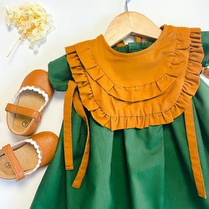May include: A green dress with a brown ruffled bib and matching ties. The dress is hanging on a wooden hanger.
