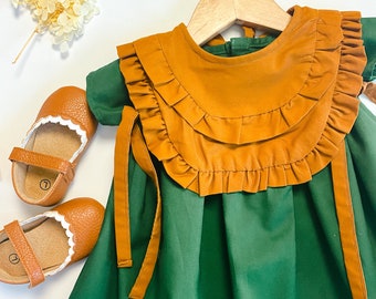 May include: A green dress with a brown ruffled bib and matching ties. The dress is hanging on a wooden hanger.