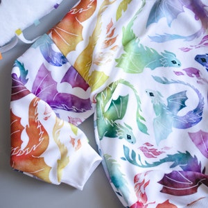 Puede incluir: Sudadera blanca con estampado de acuarela de dragones coloridos. Los dragones son en tonos de naranja, amarillo, verde, azul y morado.